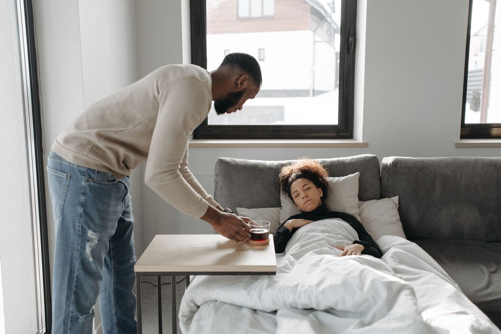 A man bringing a cup of tea to a woman resting under a blanket on the sofa