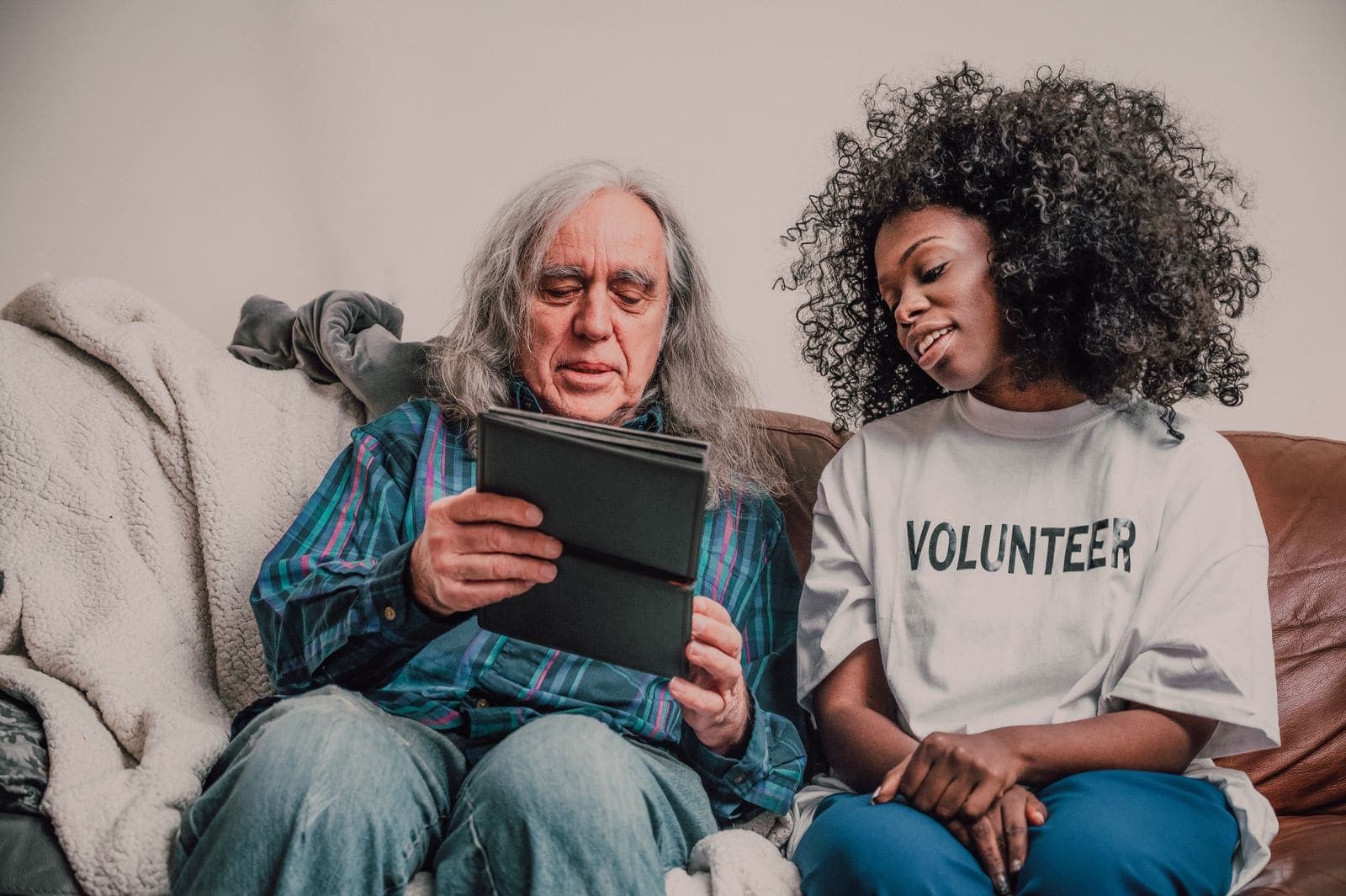 A young woman in a volunteer T-shirt helping an older man use a tablet on his sofa